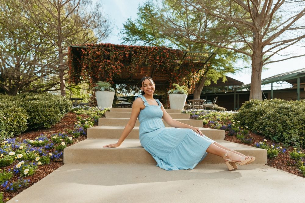 Woman in light blue dress sitting on garden steps surrounded by colorful flowers and lush greenery.