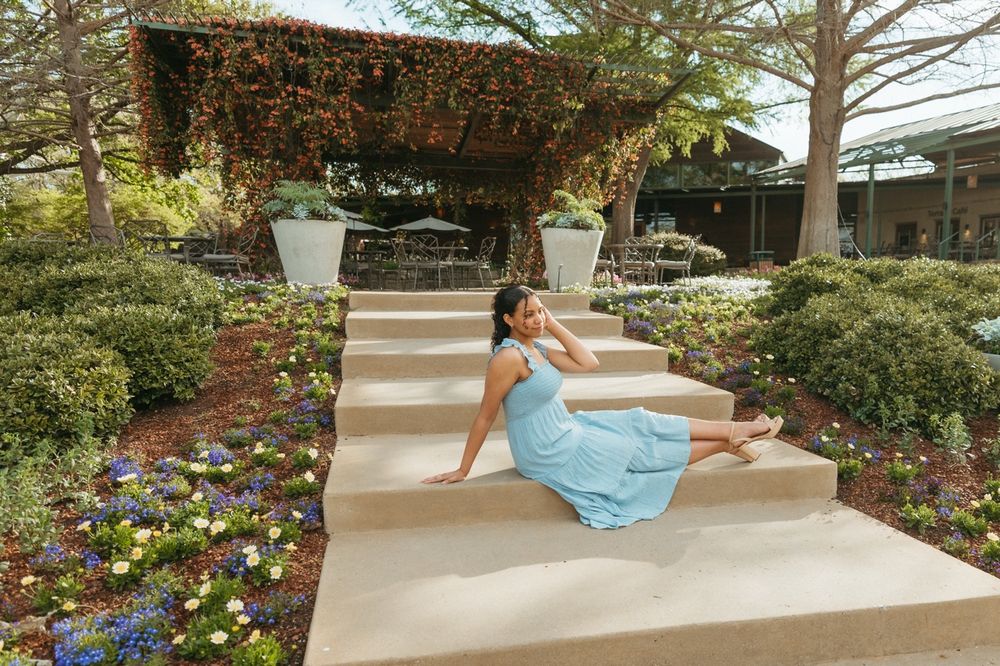 Pregnant woman in light blue dress sitting on garden steps surrounded by colorful flowers and lush greenery.