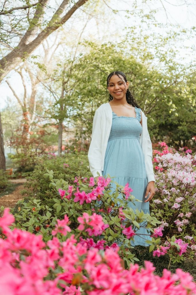 Smiling pregnant woman in blue maxi dress and white cardigan standing among vibrant pink azalea blooms.