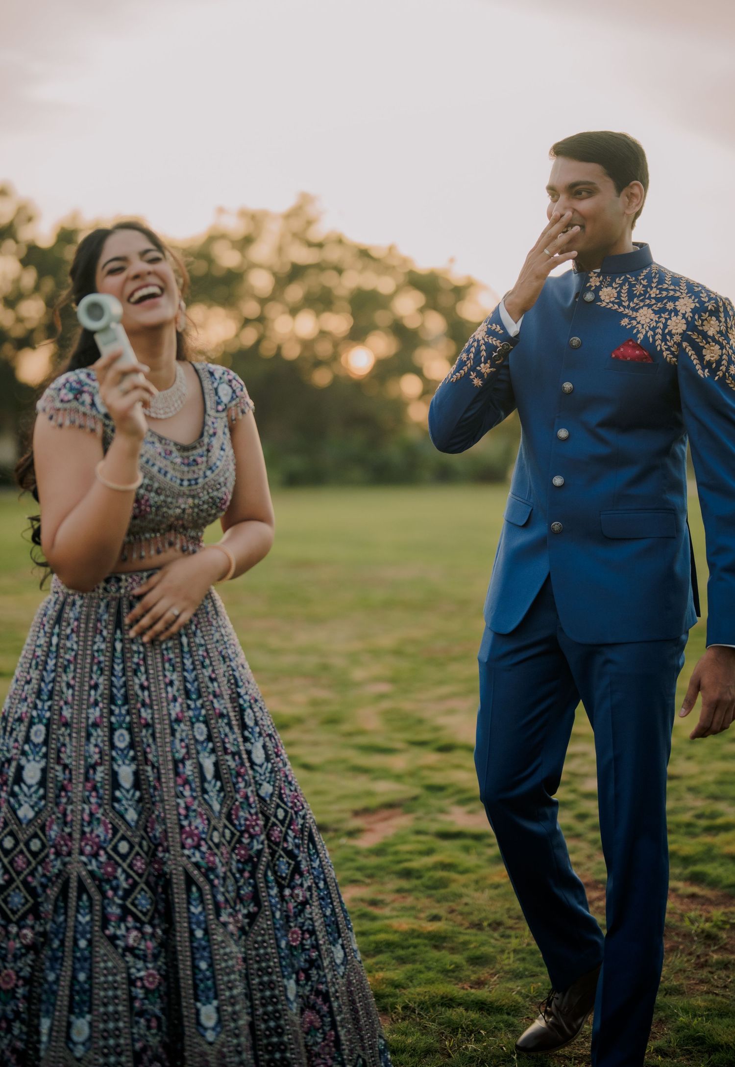 Laughing Indian couple in blue lehenga and royal blue sherwani enjoy a joyful outdoor pre-wedding photoshoot.