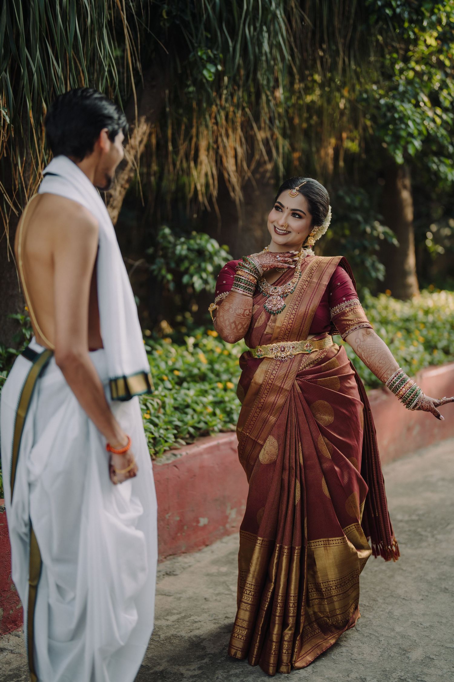 South Indian bride in traditional maroon silk saree with gold border meeting groom in white dhoti outdoors.