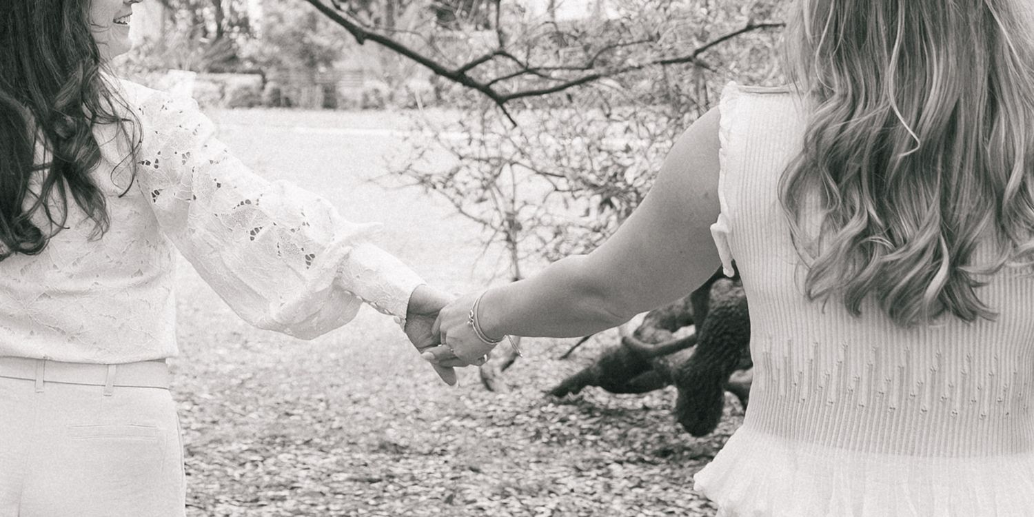 Two women holding hands in a black and white outdoor photo, wearing white dresses among autumn trees.