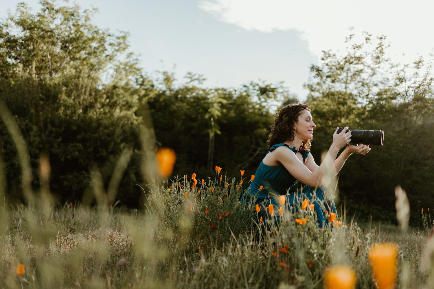 Woman photographing wildflowers in a lush garden at golden hour, surrounded by orange poppies and green foliage.