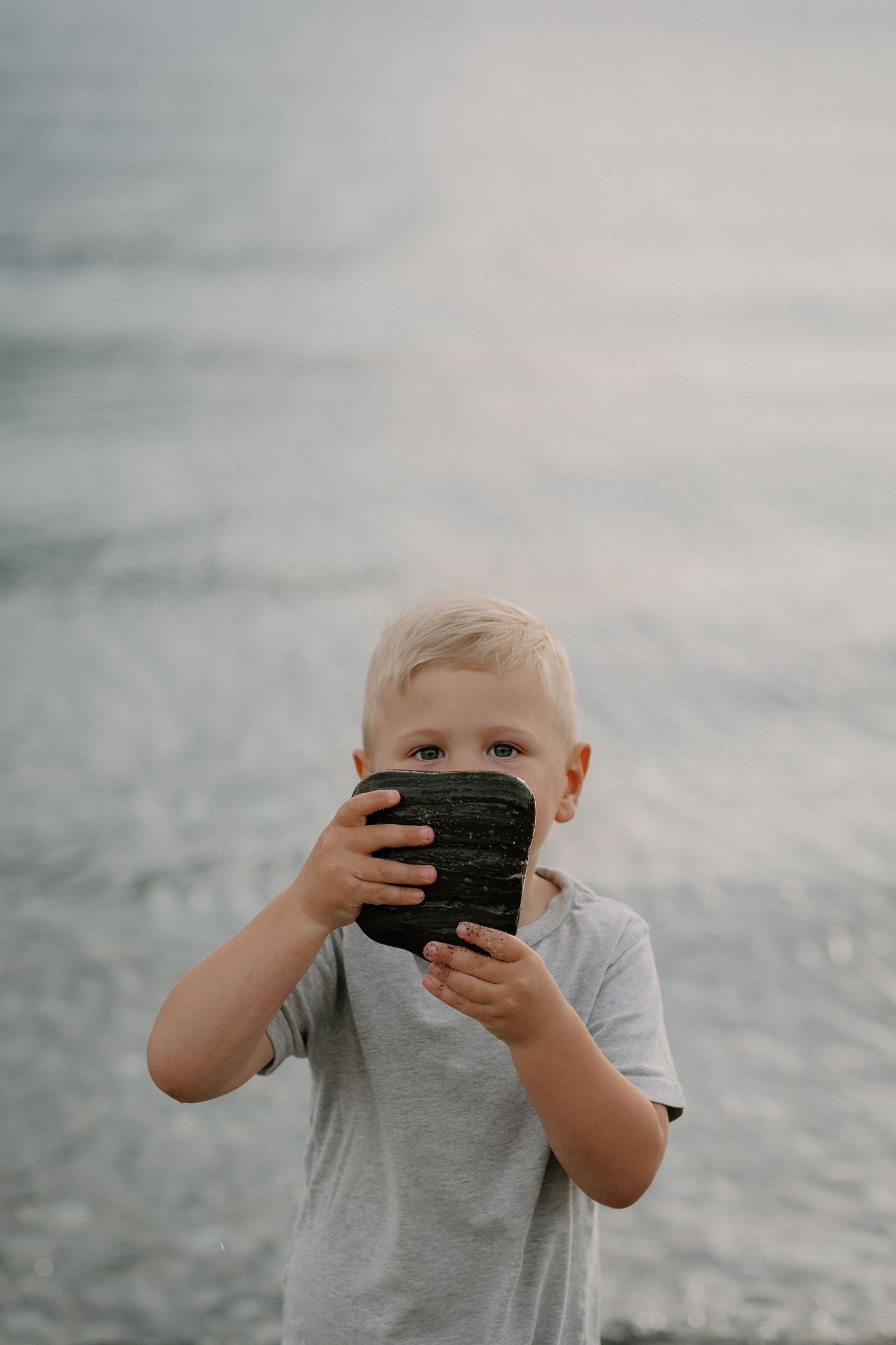 Young blonde boy holding a dark rock up to his face near a shimmering ocean shoreline at Homestead Bay in Queenstown during a family session