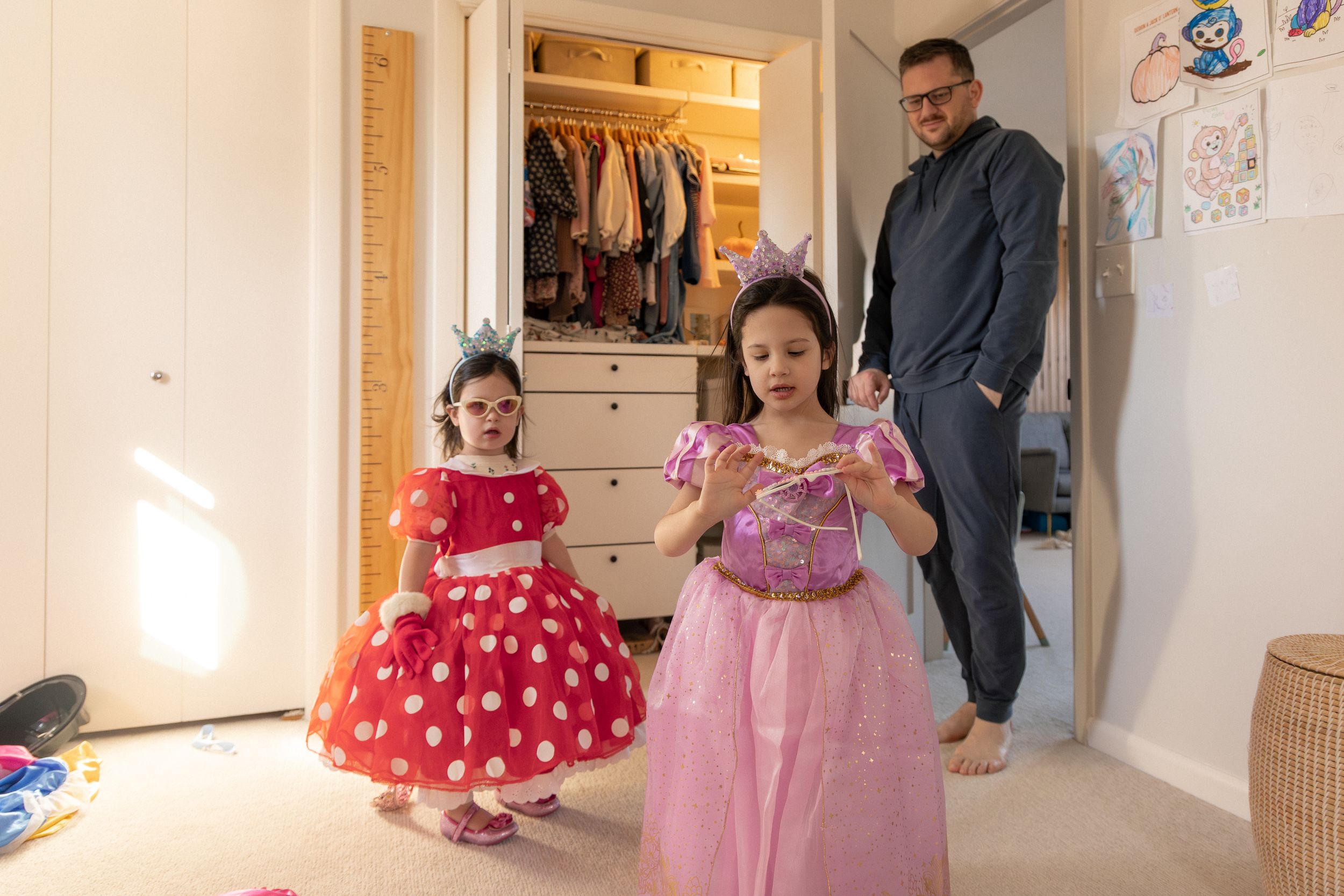 Two children wearing princess dresses play dress-up while an adult watches from the doorway of a bedroom.