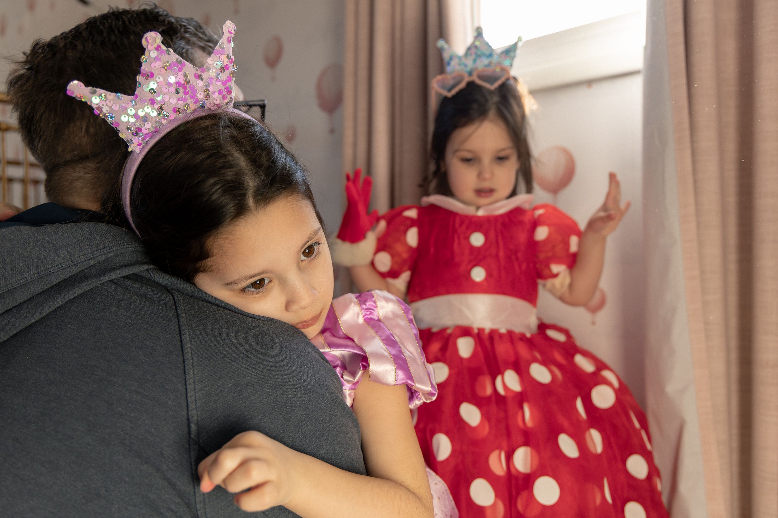 Two children in princess costumes with crowns relax on a couch, one wearing a red polka dot dress.