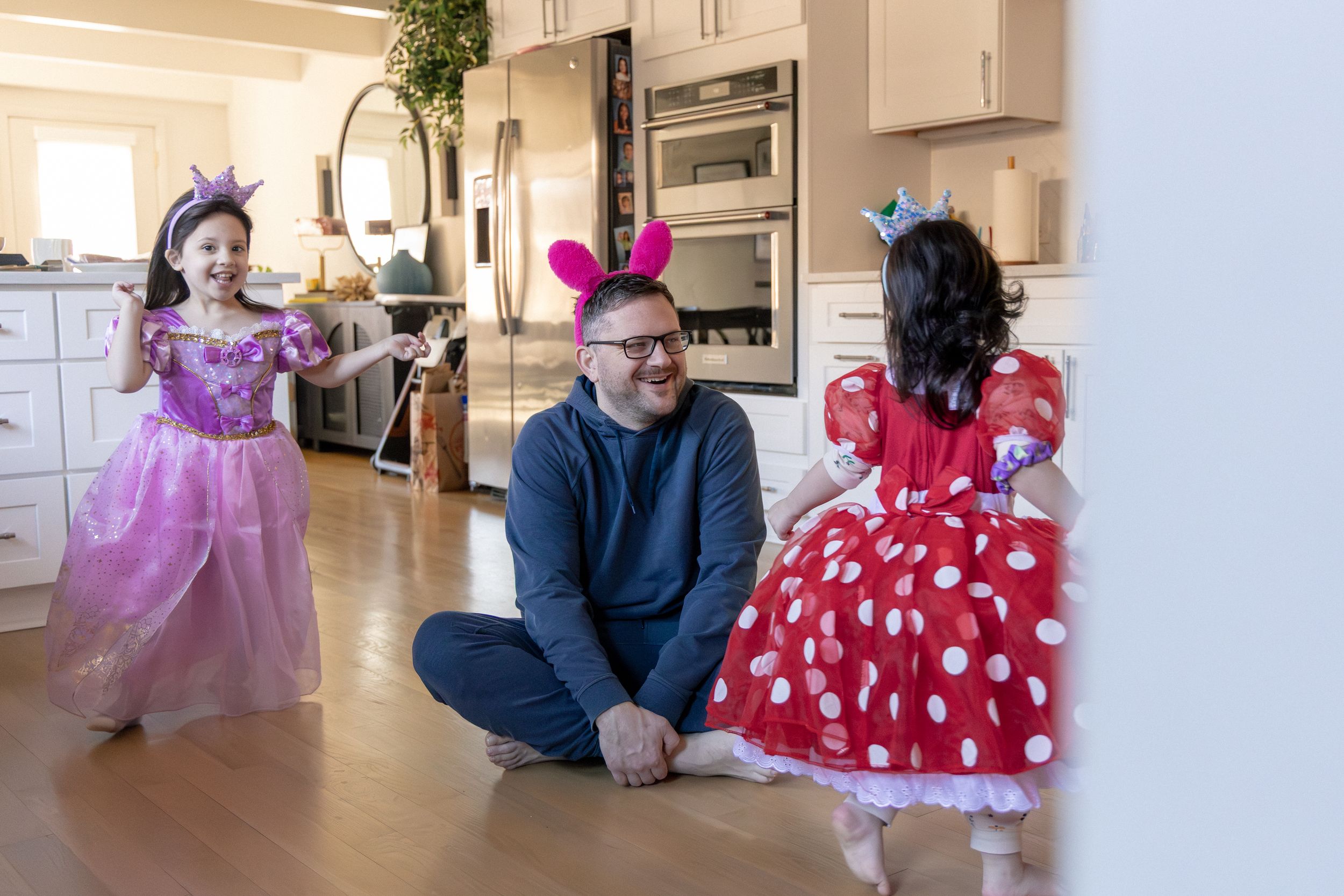 Children in princess costumes twirl and dance in a bright modern kitchen while an adult sits on the floor.