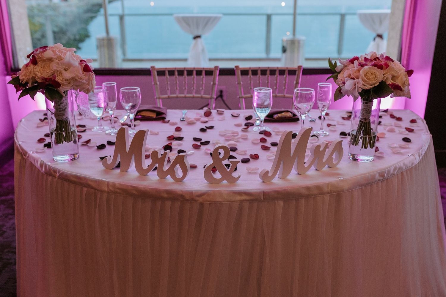 Wedding reception decor featuring elegant white tablecloths and pink uplighting in a banquet hall setting.