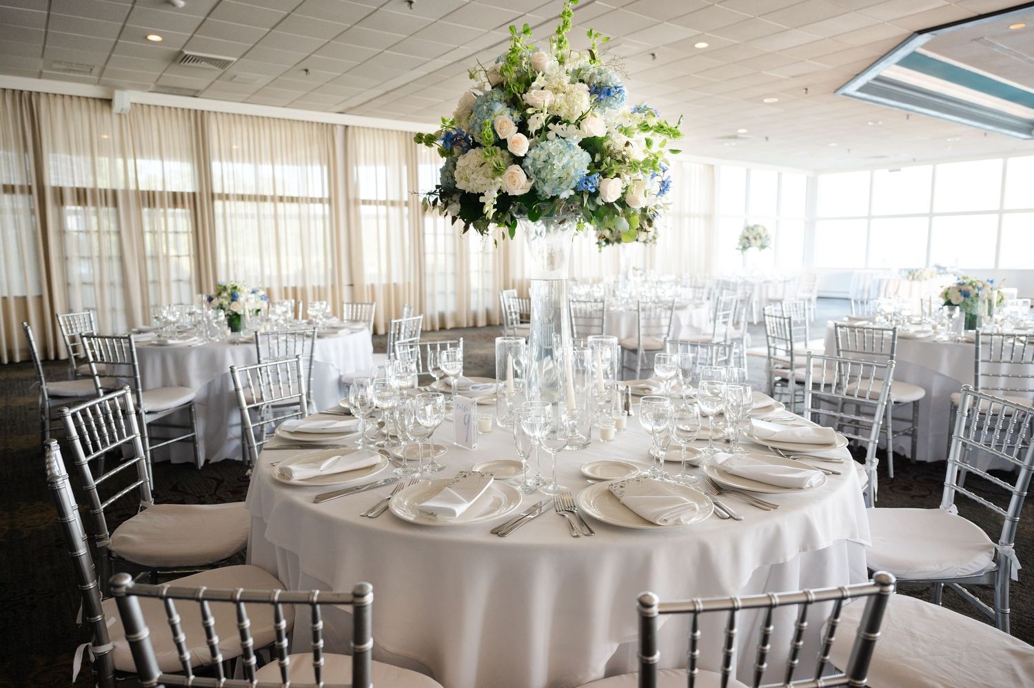 Elegant wedding reception with round tables, silver chairs, white linens, and tall floral centerpieces with blue hydrangeas in Mamaroneck.