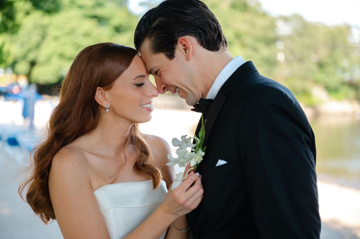 Bride and groom in wedding attire sharing an intimate moment outdoors, touching foreheads while holding a white boutonniere.