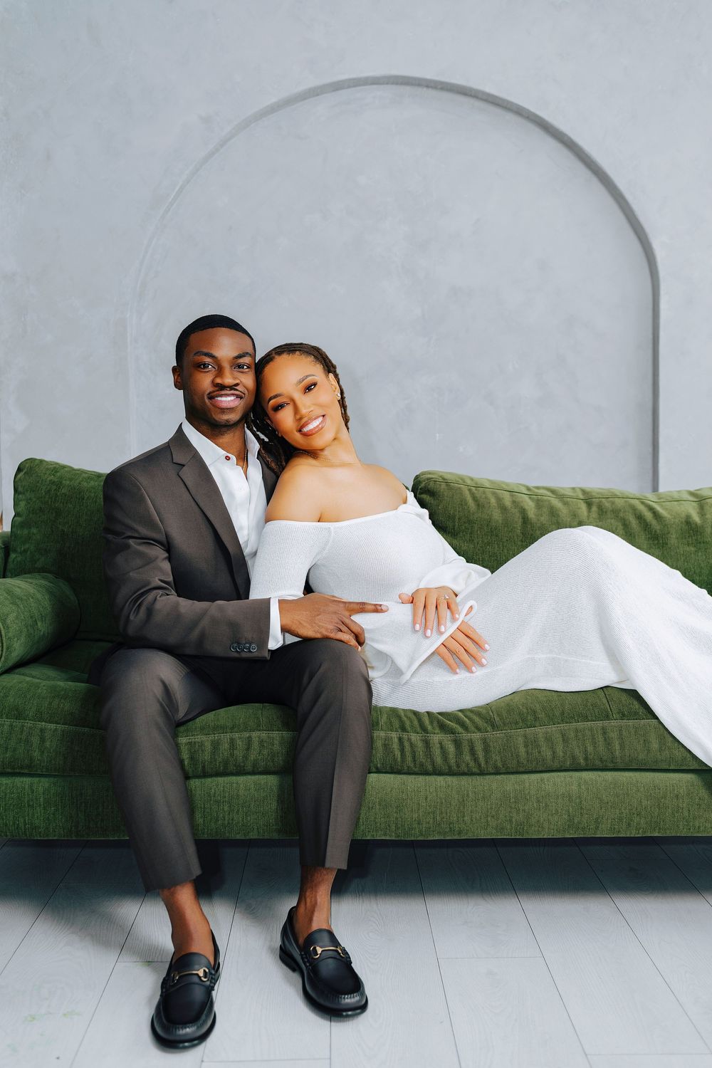 Elegant Black couple posing on green velvet sofa, man in gray suit, woman in white off-shoulder gown.