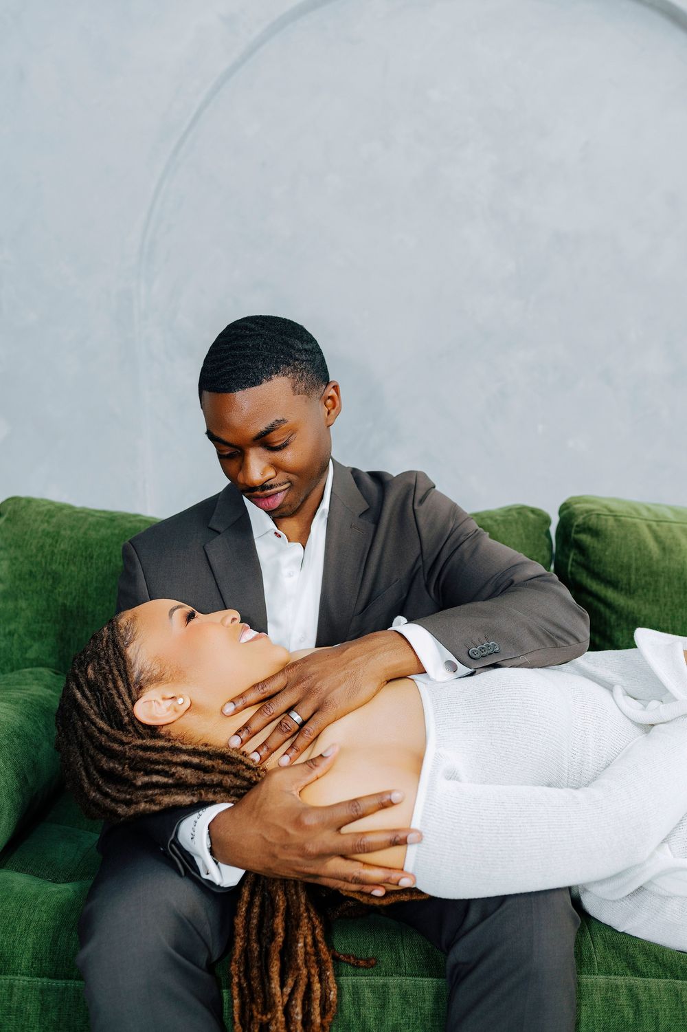 A couple shares an intimate moment on a green velvet sofa, man in gray suit cradling woman in white dress.