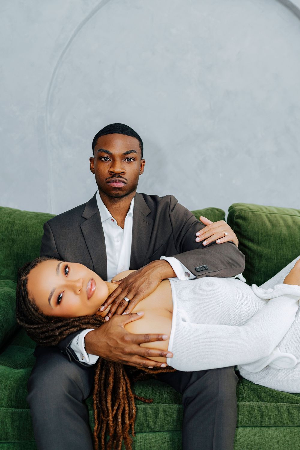 A Black couple poses on a green velvet sofa, man in dark suit, woman in white with braids resting on his lap.