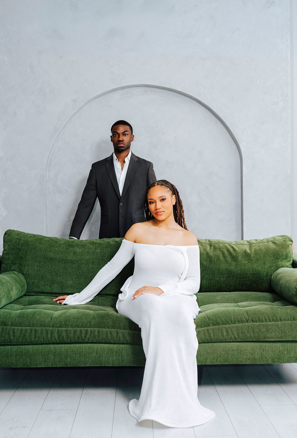 Elegant couple in formal attire posing in a studio with a green velvet sofa and arched wall backdrop.