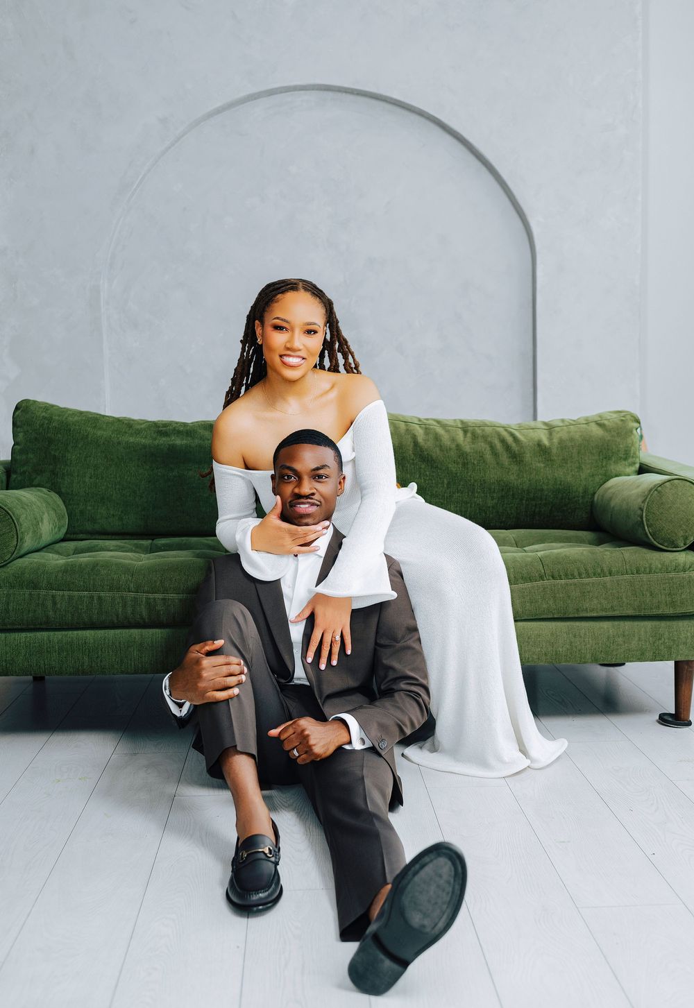 Black bride in white off-shoulder gown embraces groom in gray suit before green velvet sofa.