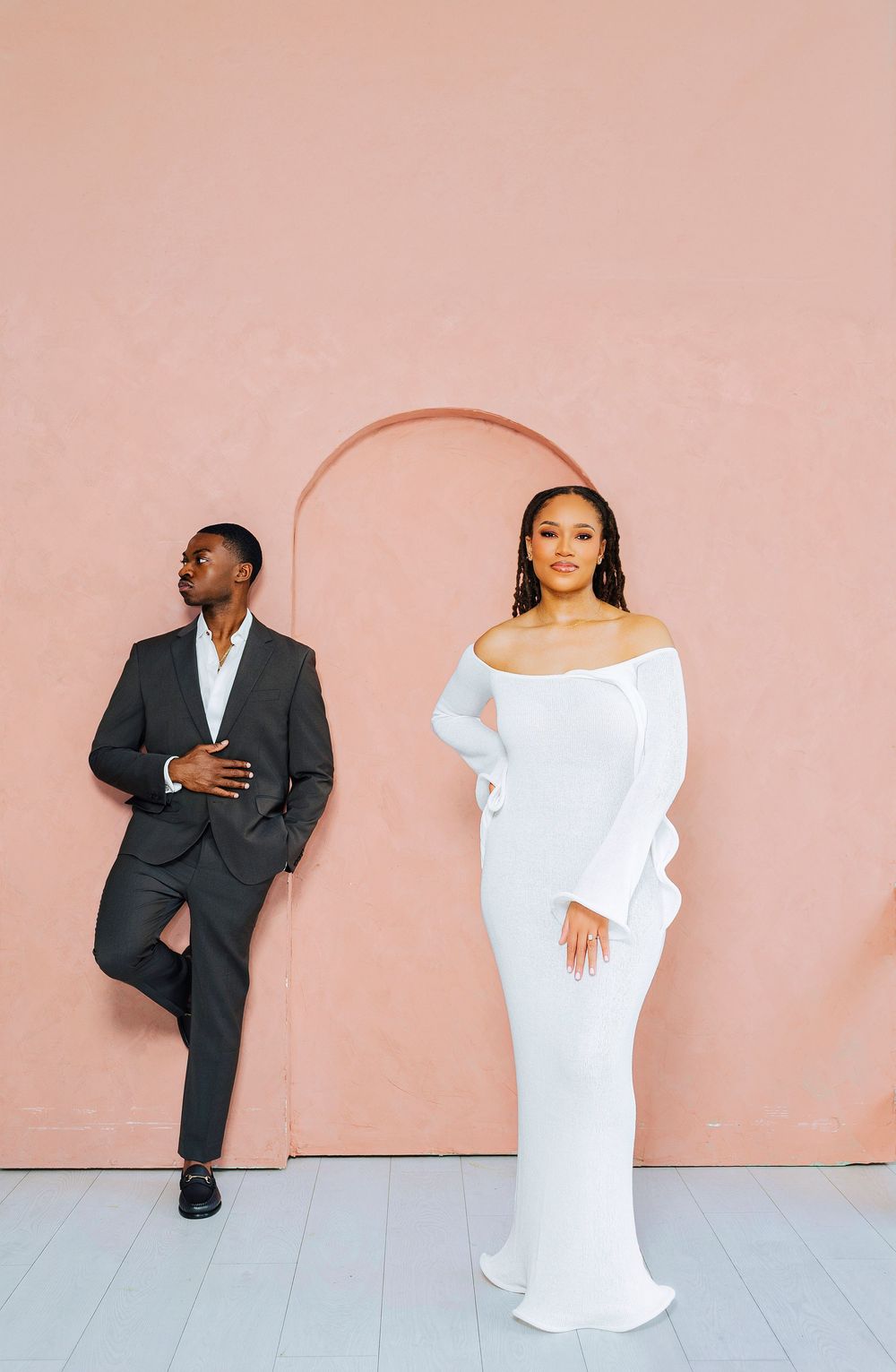 Elegant couple posing against a pink wall, man in dark suit and woman in white off-shoulder gown.