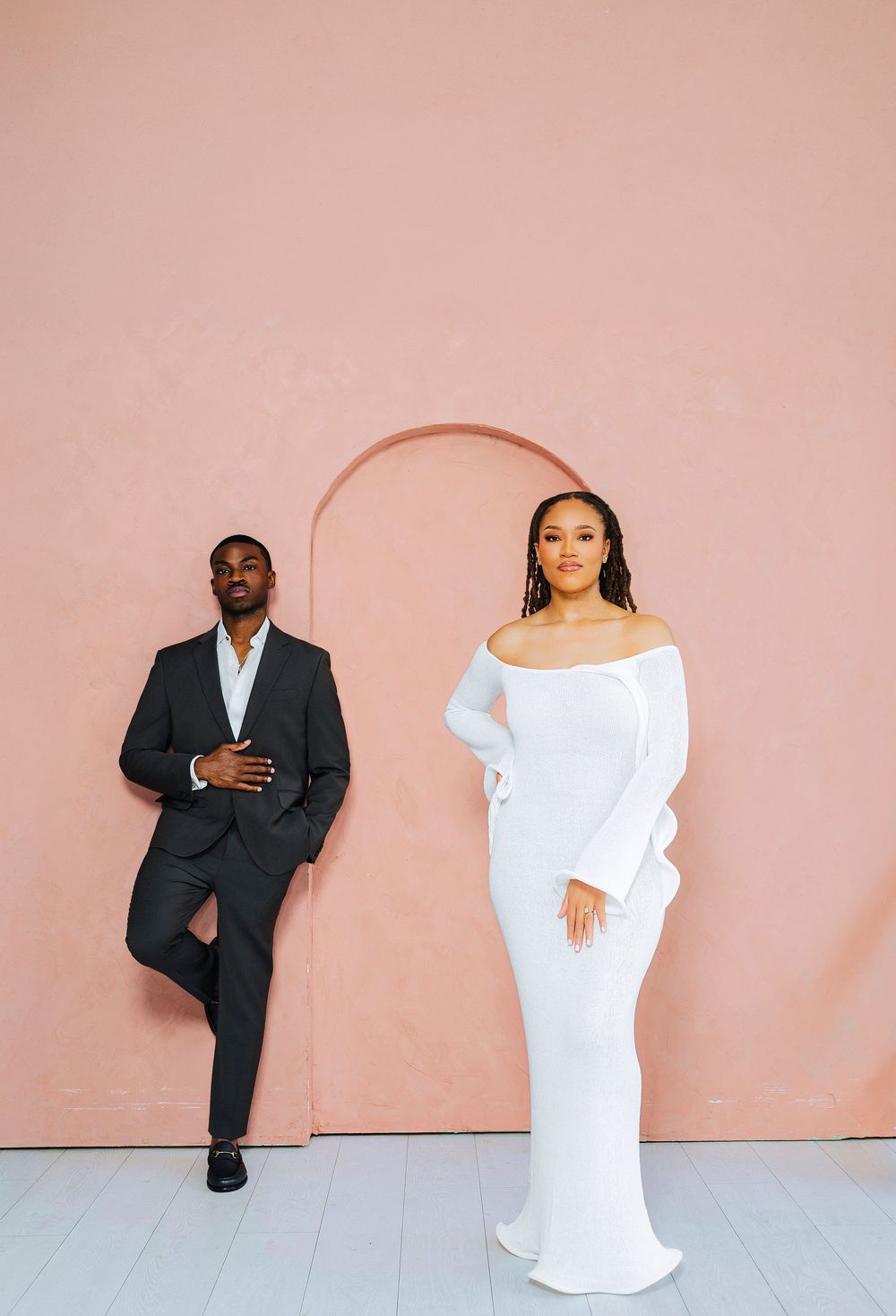 An elegant Black couple poses before a pink arched wall, man in dark suit, woman in a white off-shoulder gown.