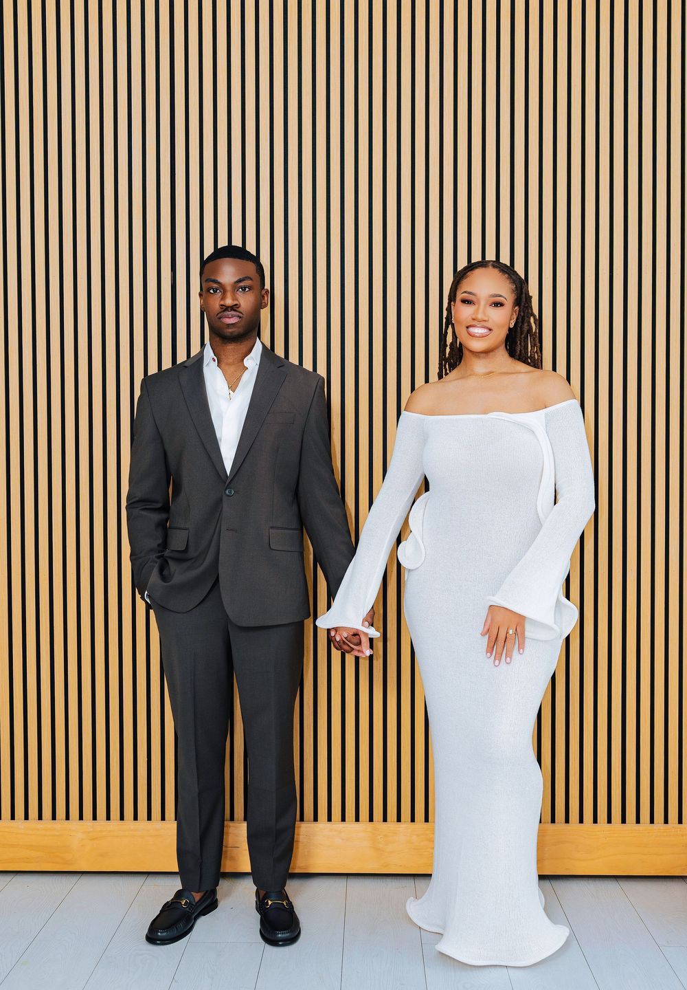 Elegant couple posing hand-in-hand, man in dark suit and woman in white off-shoulder gown, against wood panel wall.