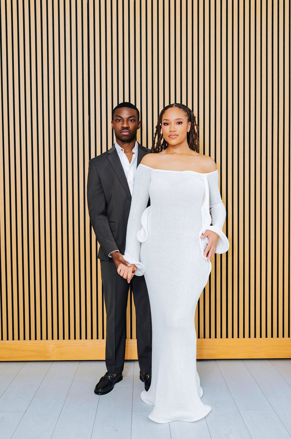 Stylish Black couple holding hands before wooden slat wall, bride in fitted white gown, groom in gray suit.