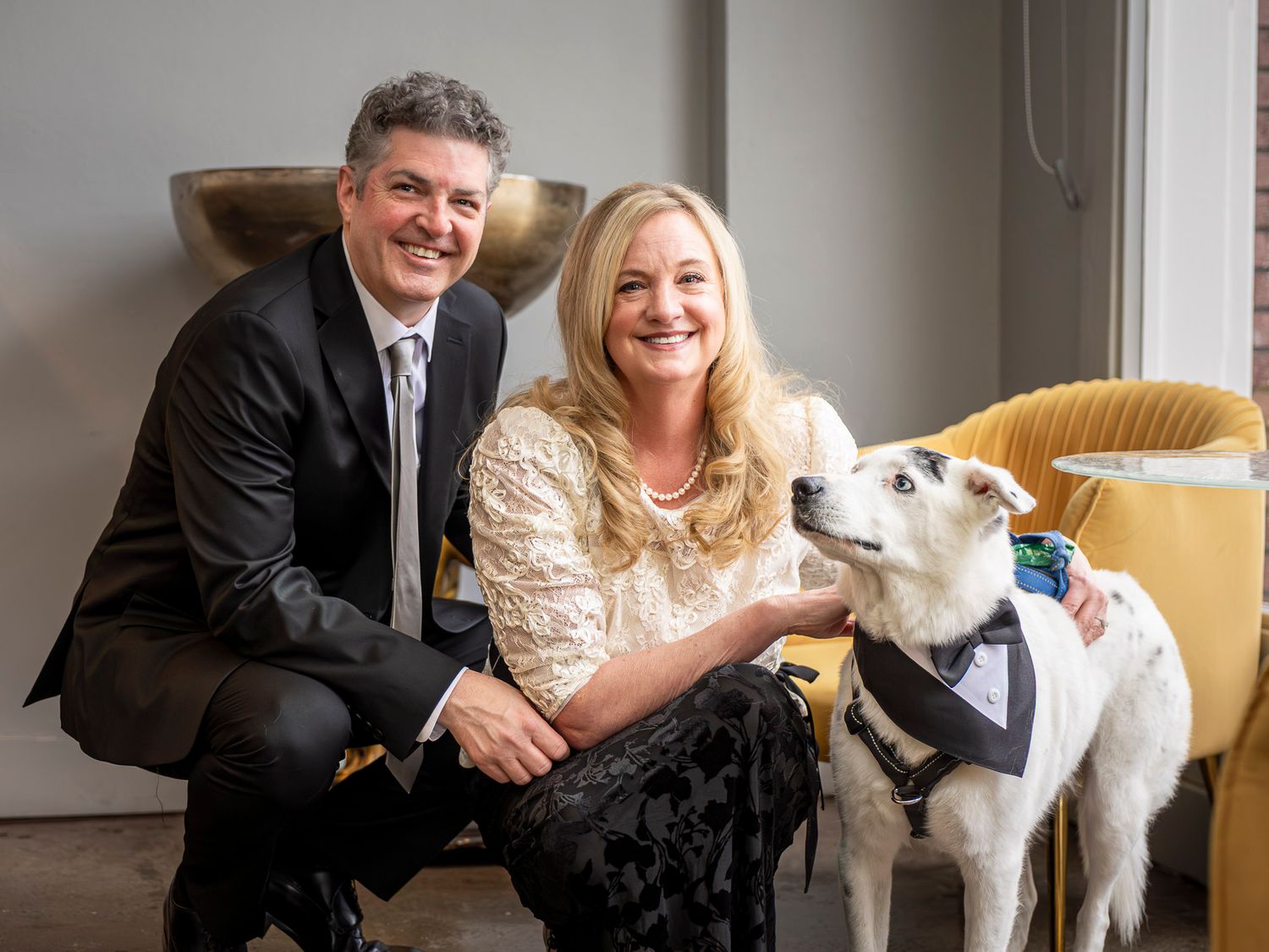 Couple in formal attire smiling with a white dog wearing a tuxedo bandana at an elegant indoor setting.