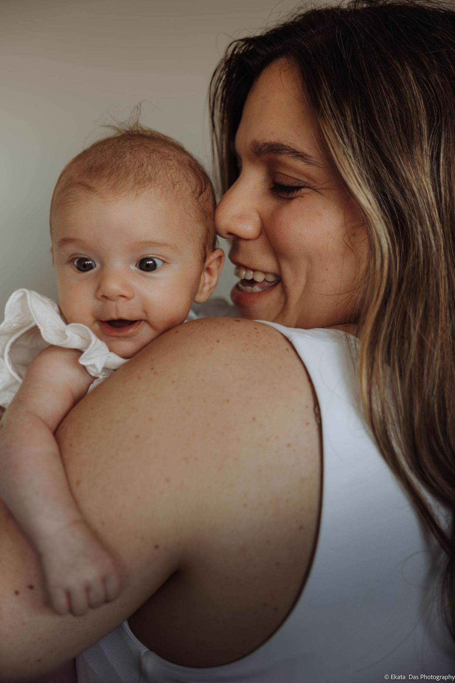 A smiling woman holds a happy baby in a white onesie, sharing a joyful moment together.