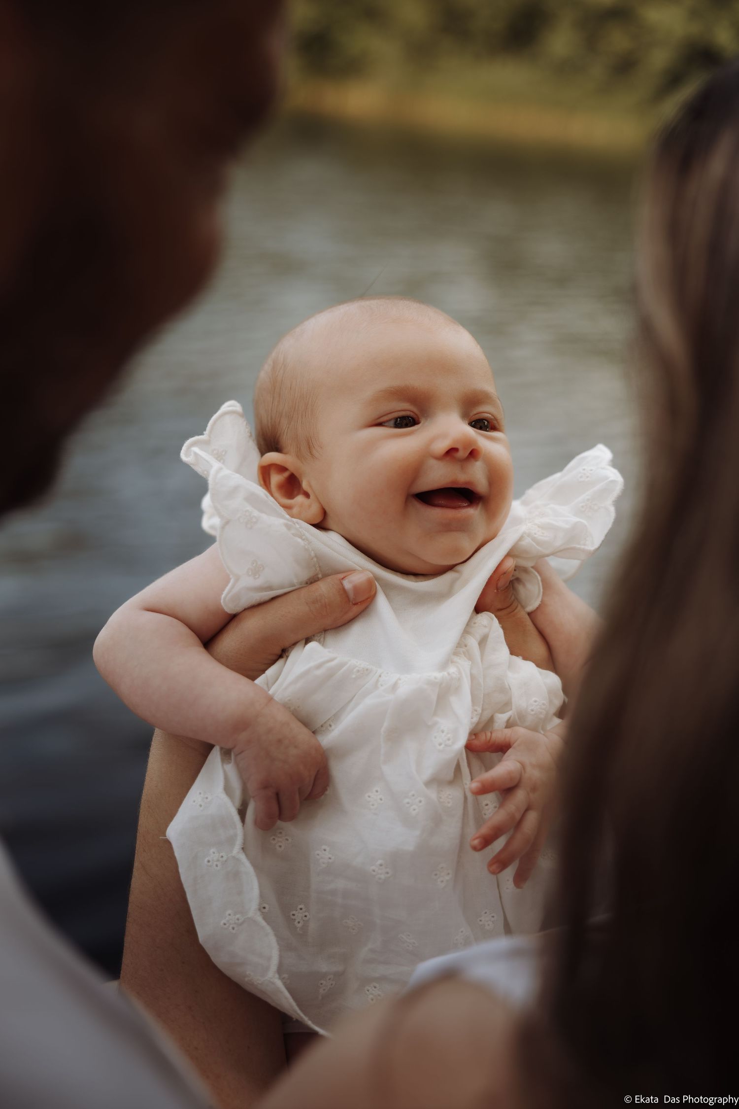 Smiling baby in white dress held by parents outdoors near water during a family photo session.