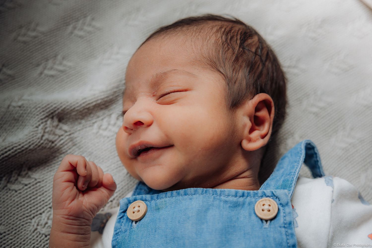Smiling newborn baby with eyes closed lying on a soft gray blanket, wearing a blue denim overalls outfit with wooden buttons.