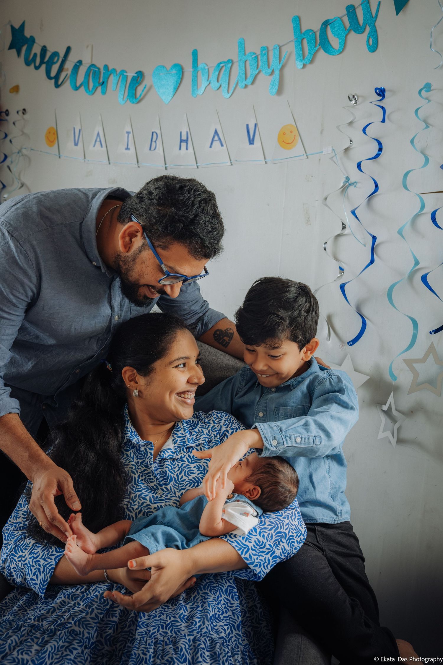 Smiling parents and their young son gather around a newborn baby under a blue welcome baby banner during a family photoshoot.