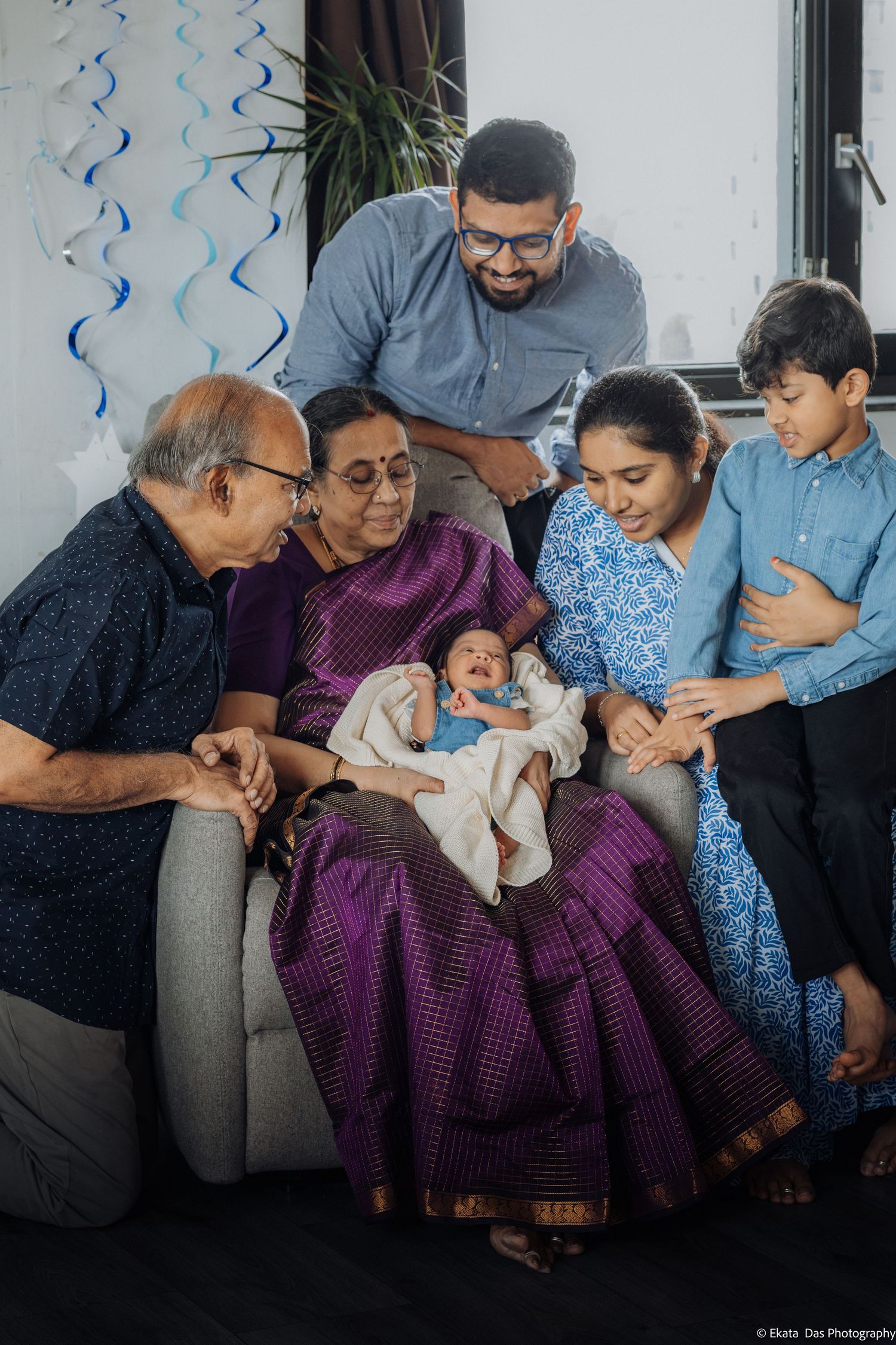 Multigenerational family gathered around a newborn baby held by a grandmother in a purple sari, with a smiling boy and adults nearby.