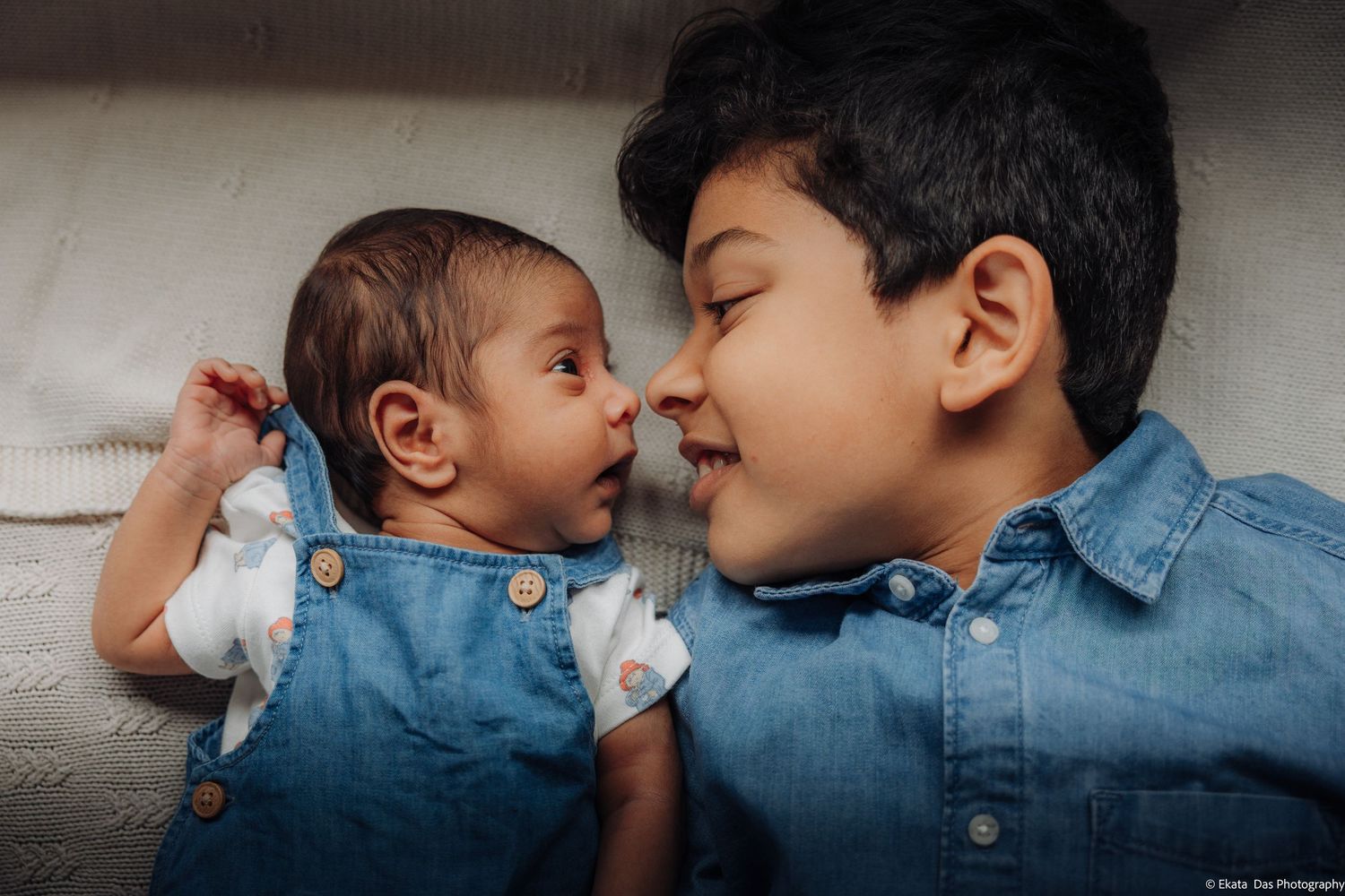 Older brother in a blue denim shirt lies nose to nose with his newborn sibling in a denim overall on a cozy cream blanket.