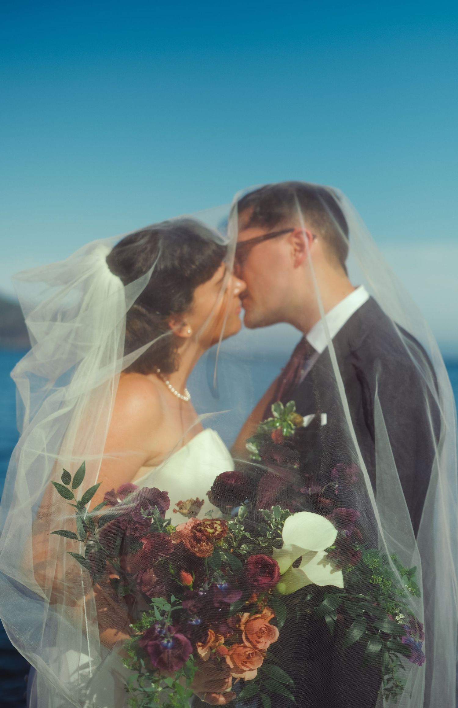 Bride and groom kissing under a veil outdoors, bride holding a lush bouquet of burgundy and white flowers with a blue sky backdrop.