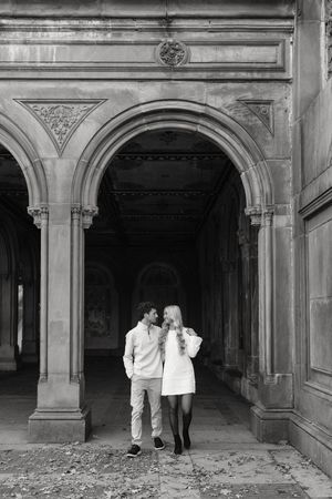 Black and white photo of a couple sharing a romantic moment under ornate stone arches at Bethesda Terrace, Central Park.