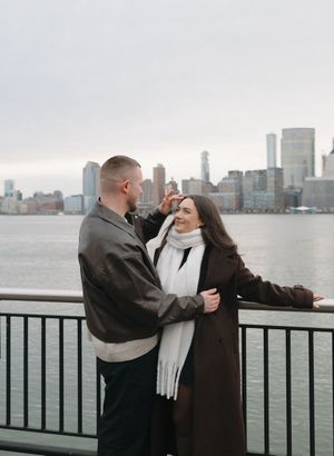 Couple sharing a romantic moment by a waterfront railing with a city skyline backdrop on a cloudy day.