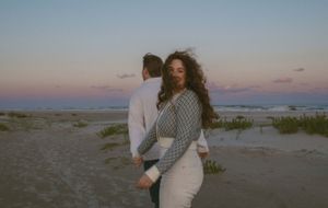 Couple holding hands on a sandy beach at sunset, woman in white dress with curly hair smiling over her shoulder.