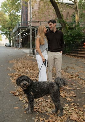 A nyc couple smiles at each other on a leaf-covered sidewalk while walking their large black doodle dog on a leash.