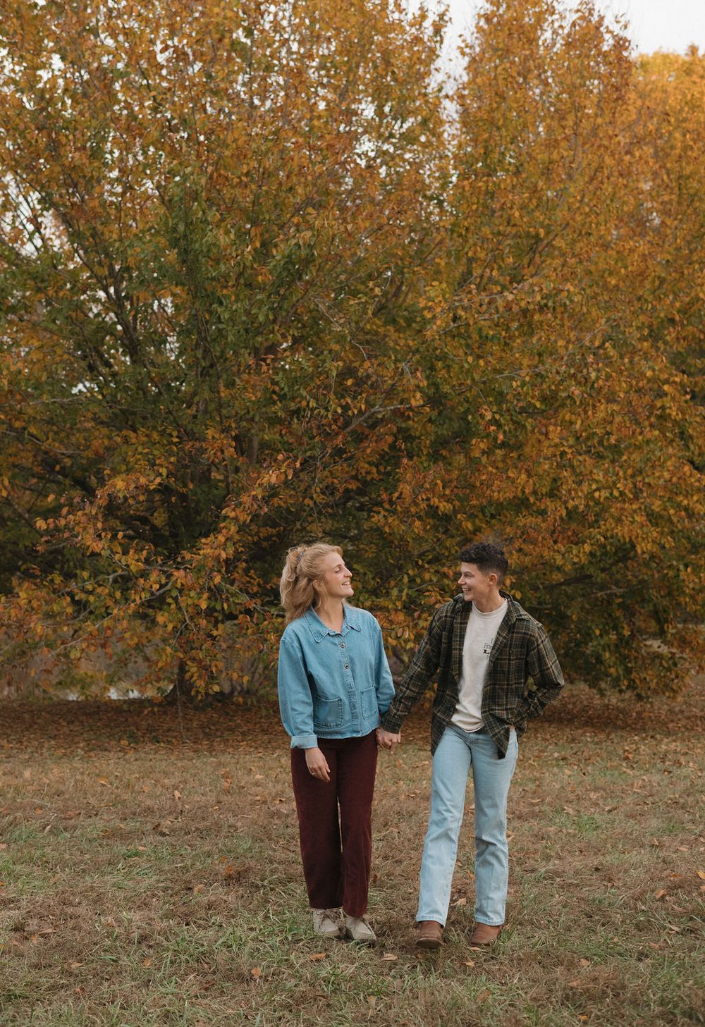Two people hold hands in a field surrounded by autumn trees with golden foliage during a fall outdoor photoshoot.