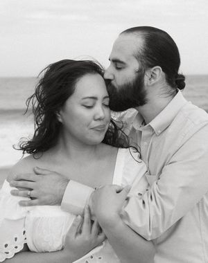 Black and white beach photo of a bearded man kissing a woman on the head while embracing her tenderly.