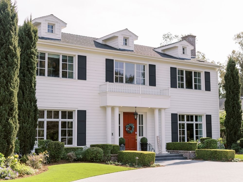 Elegant white colonial style house with red door, tall windows, and manicured cypress trees in the landscaping.