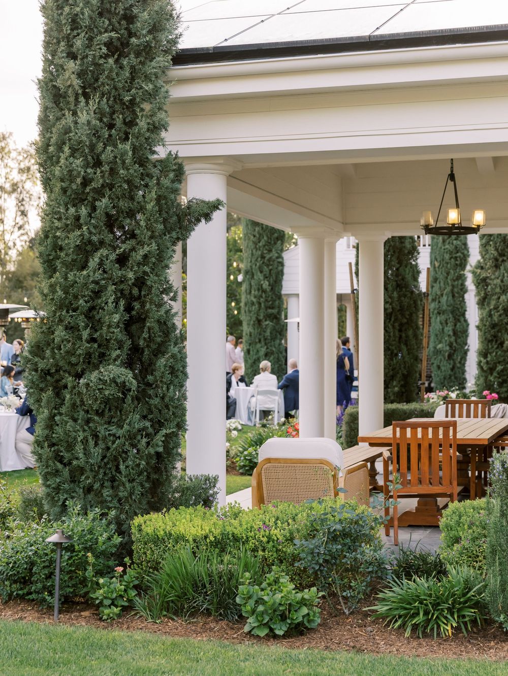 White columns and wooden benches adorn an elegant outdoor porch area with lush greenery and cypress trees.