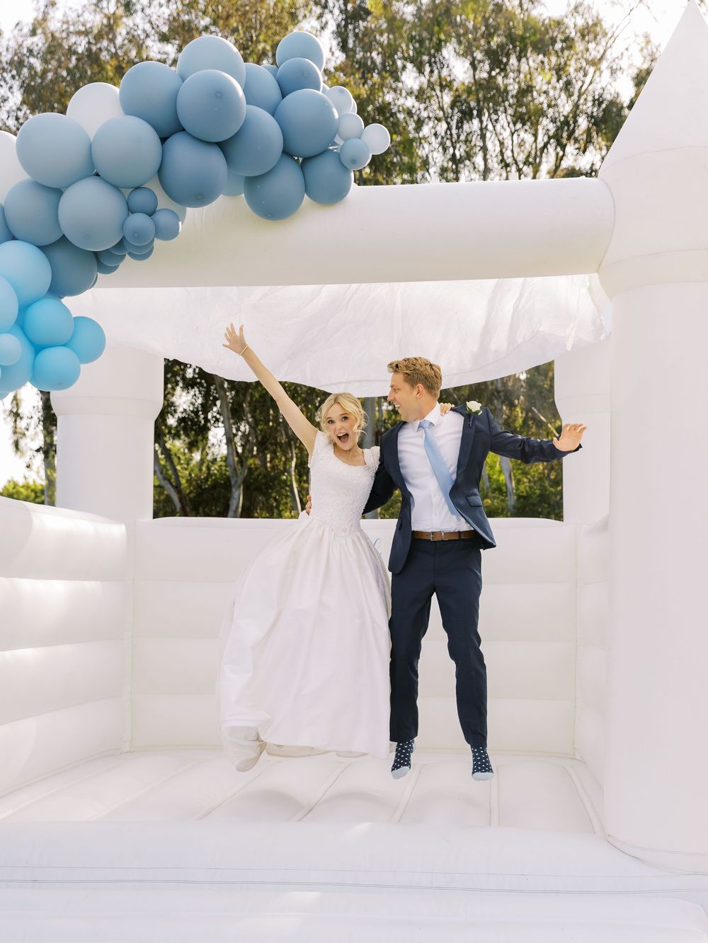 White bounce house decorated with blue balloon arch creates a playful wedding photo backdrop at outdoor celebration.
