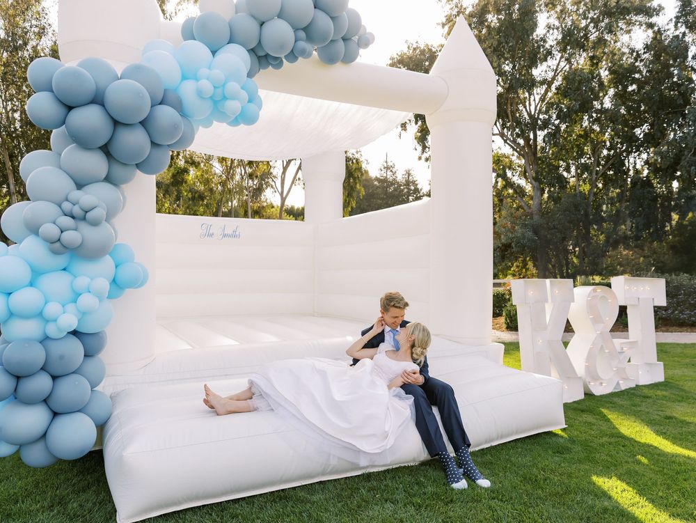 White bounce house with blue balloon decorations set up on a sunny lawn.