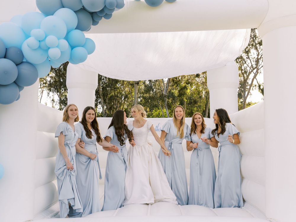 Wedding party wearing coordinated light blue dresses standing together under blue balloon arch.