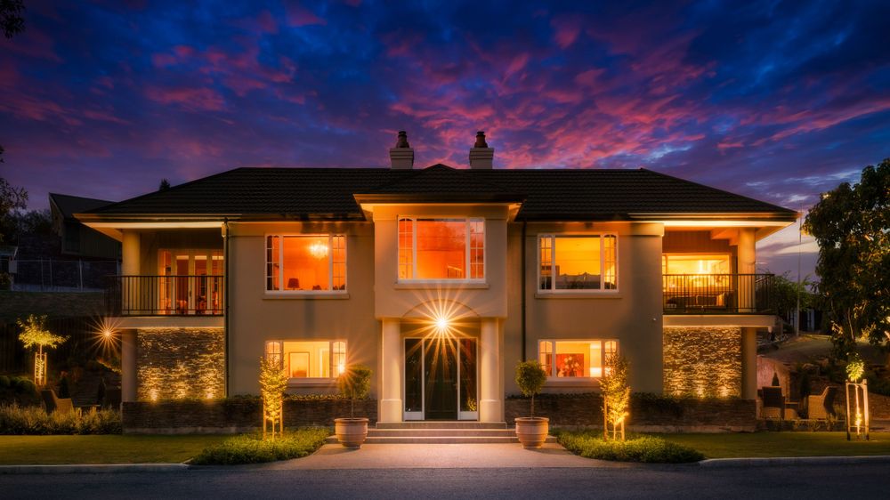 Luxurious modern home exterior illuminated at dusk with warm glowing windows and landscaping lights.