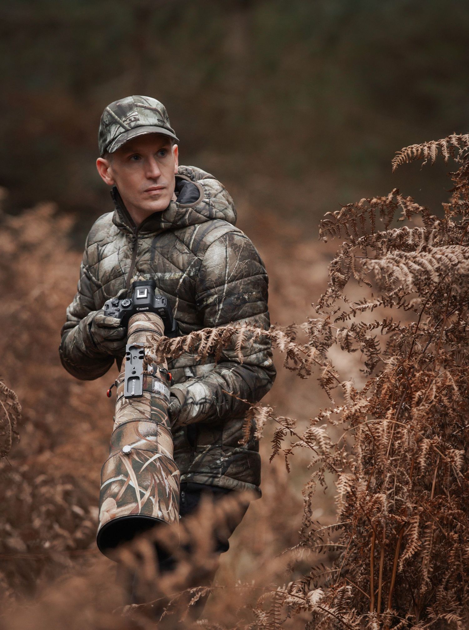 Wildlife photographer in camouflage gear positions telephoto lens among autumn foliage in natural setting.