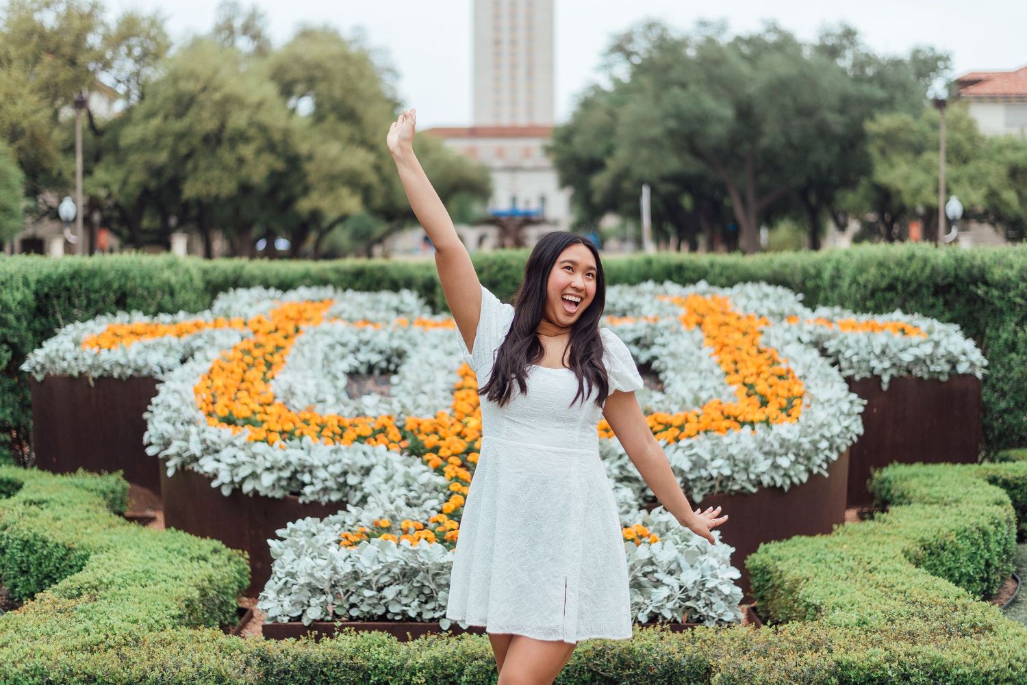 A joyful figure in a white dress poses in front of a butterfly-shaped flower garden at the University of Texas campus.