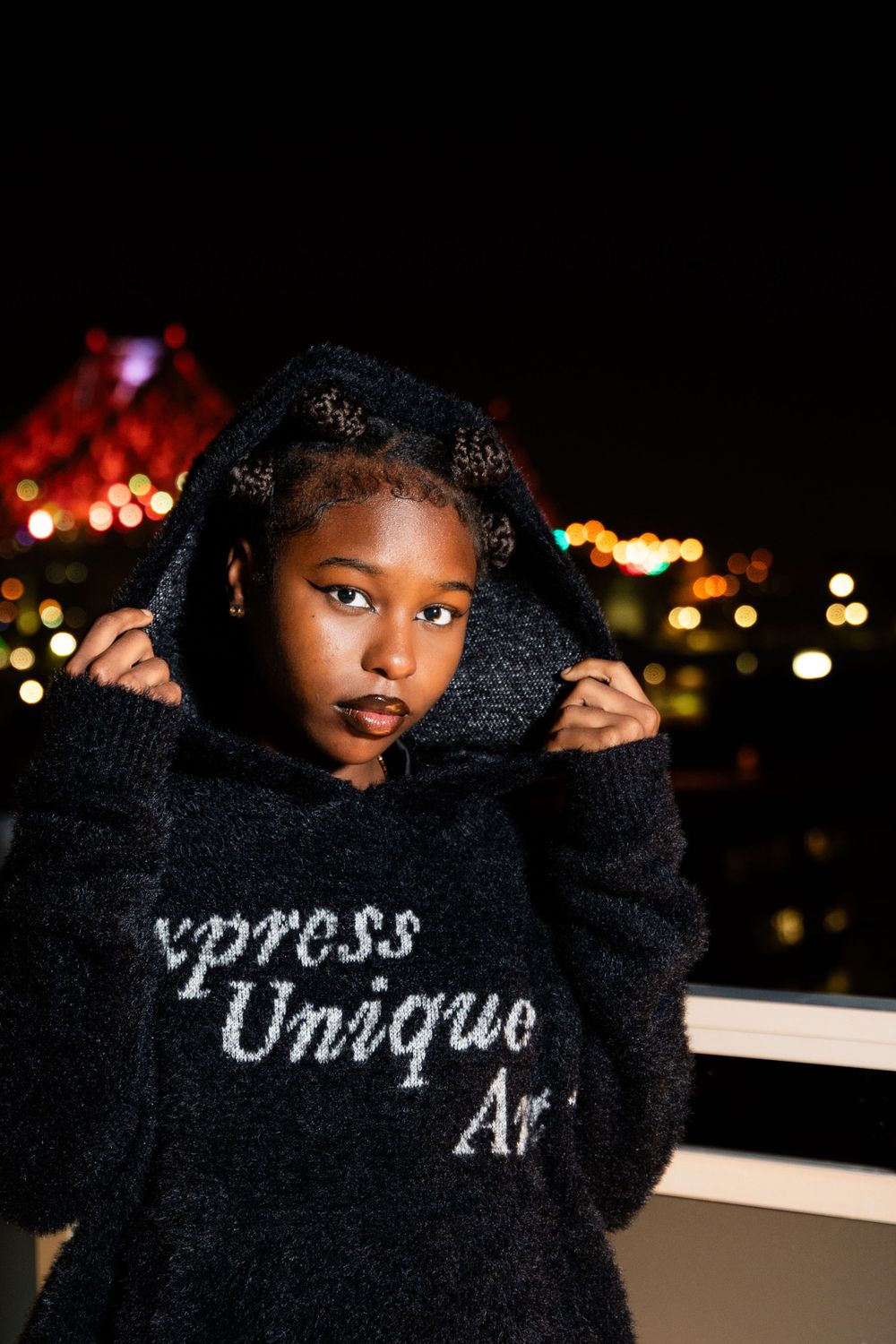 Nighttime portrait in black fuzzy hoodie with 'Express Unique AF' text against city lights.