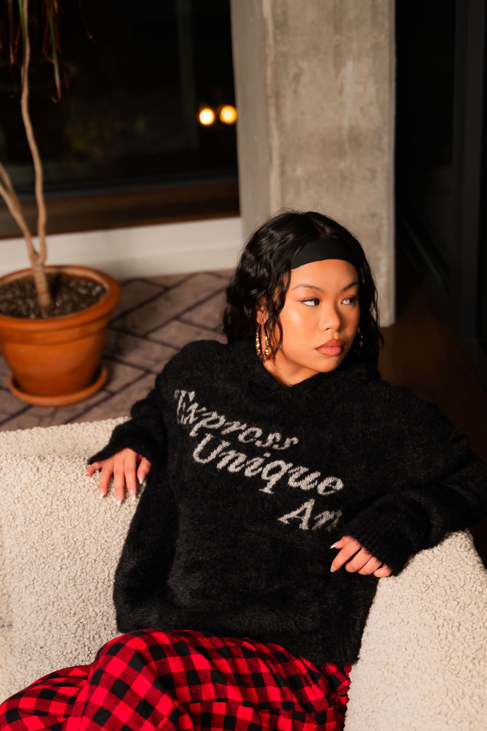 Casual indoor portrait with black sweater and red checkered pants near window.