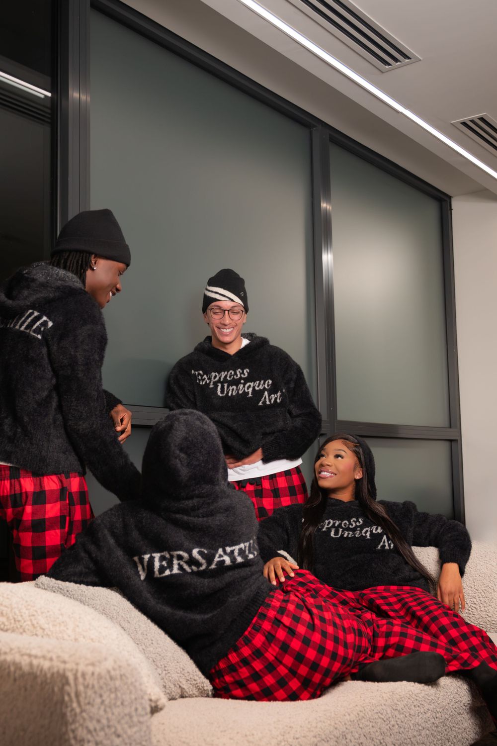 Friends wearing matching black hoodies with red plaid pants relaxing on a couch during a casual photoshoot.