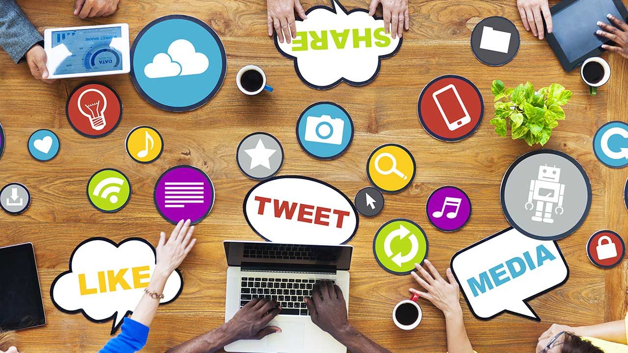 Overhead view of hands using devices with social media icons and chat bubbles on wooden table.