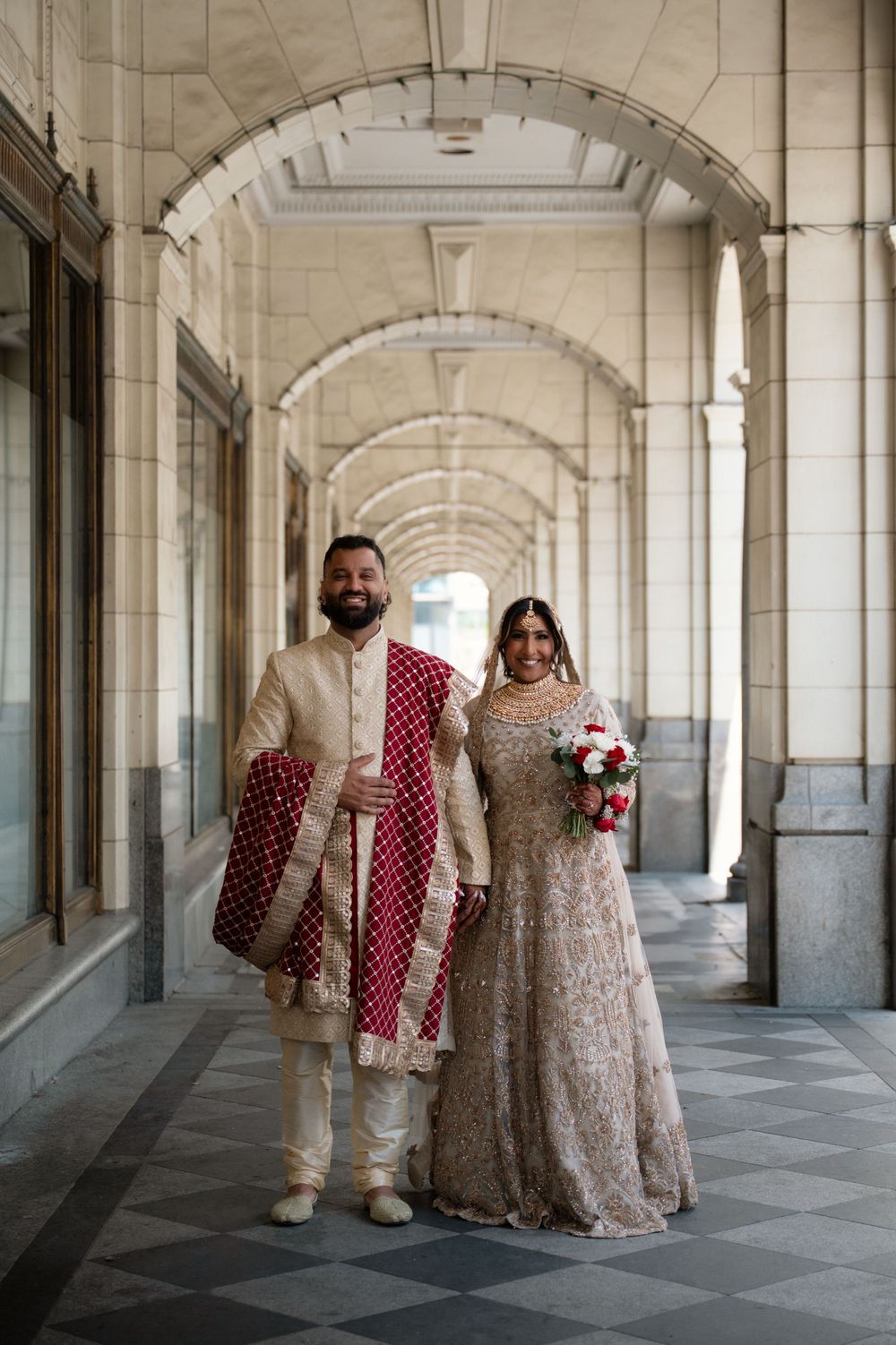 Couple in traditional South Asian wedding attire stands in an elegant colonnade with arched ceilings.